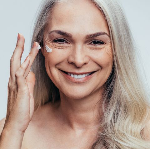 smiling mid adult caucasian woman applying anti aging cream on her face senior female woman applying moisturizer on her face against grey background