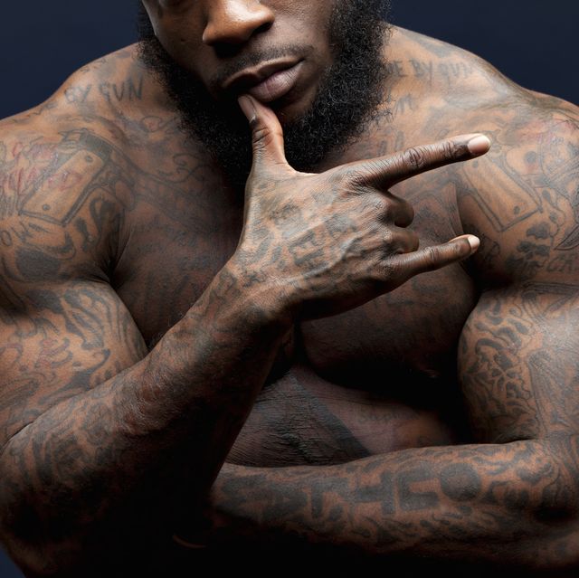 Der mit Tätowierungen bedeckte afroamerikanische Mann zeigt seine Brust-, Schulter- und Armtätowierungen von Waffen, verschiedene Phrasen und Designs auf seinem ganzen Körper, seine Hand auf seinem Gesicht