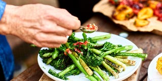 Person spooning sauce over freshly cooked green vegetable medley dish with fresh green tenderstem brococli and asparagus and red chilli dressing, serving, plating up, food preparation, healthy eating