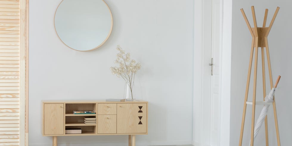 wooden hanger with white umbrella in real photo of entrance hall interior with cupboard with books and plant in glass vase and round mirror on the wall