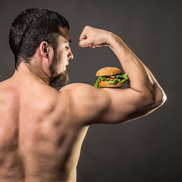 young man bodybuilder holding burger, view from behind athletic man with fast food on a gray background