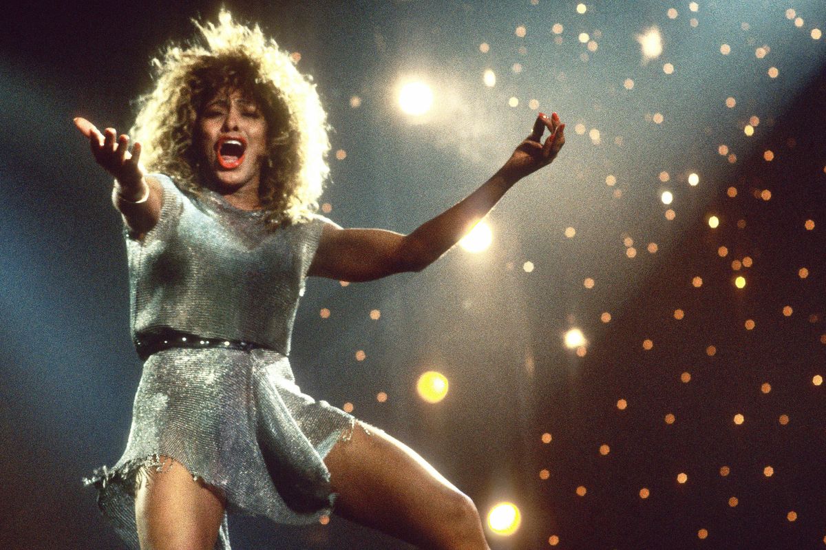 tina turner singing on stage with her arms extended, she wears a silver dress with a belt