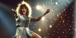 tina turner singing on stage with her arms extended, she wears a silver dress with a belt