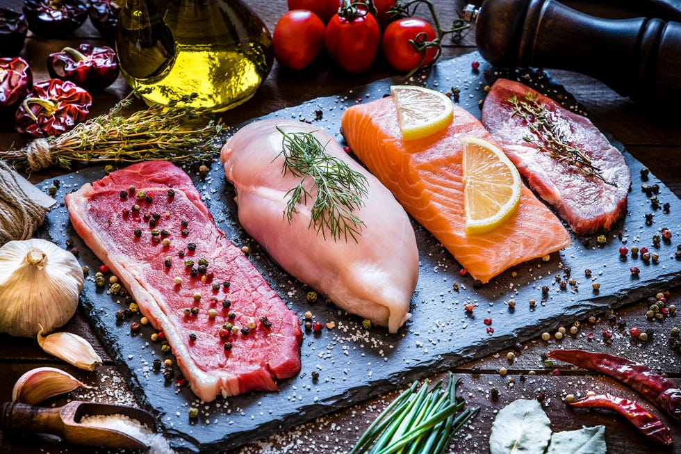 high angle view of four different types of animal protein like a raw beef steak, a raw chicken breast, a raw salmon fillet and a raw pork steak on a stone tray surrounded by condiments, spices and vegetables low key dslr photo taken with canon eos 6d mark ii and canon ef 24 105 mm f4l