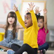 a multi ethnic group of preschool students is sitting with their legs crossed on the floor in their classroom the mixed race female teacher is sitting on the floor facing the children the happy kids are smiling and following the teacher's instructions they have their arms raised in the air