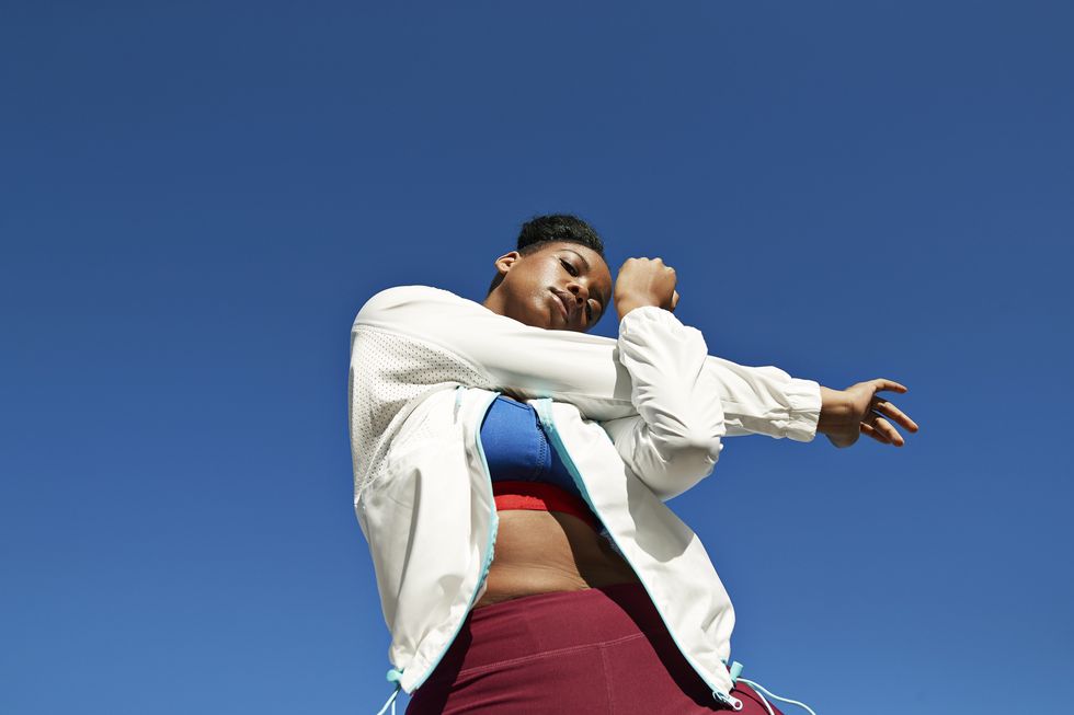 directly below of young woman wearing sports clothing while stretching arm against clear blue sky on sunny day