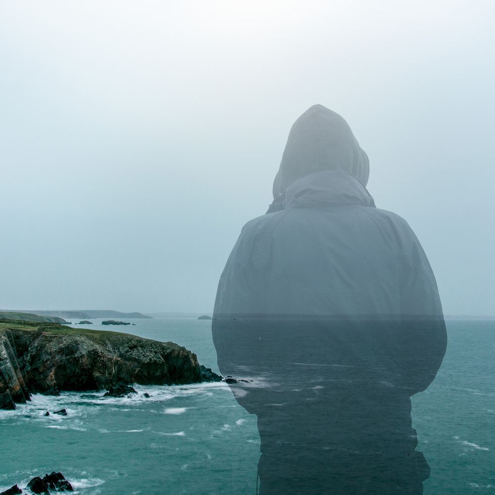 A double exposure of a hooded figure looking out at a moody rugged coast in winter.