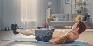 Strong Athletic Shirtless Fit Man in Grey Shorts is Doing Abdominal Exercises at Home in His Spacious and Sunny Living Room with Minimalistic Interior.