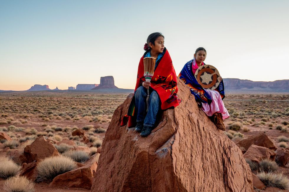 young navajo brother and sister in monument valley posing on red rocks in front of the amazing mittens rock formations in the desert at dawn