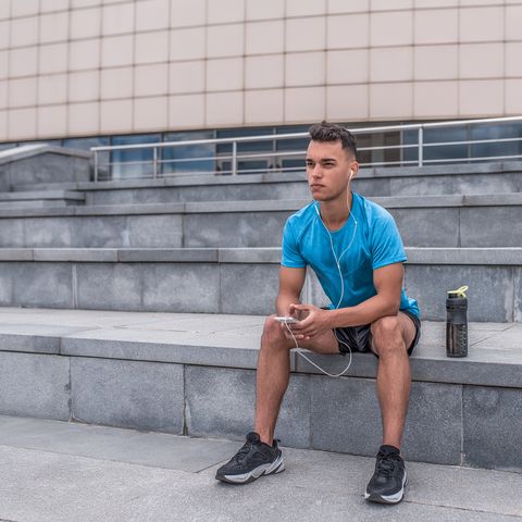 Athletic male athlete, summer city sitting hand, phone listens music headphones motivation fitness sports workout, shaker protein, morning workout. Active lifestyle young people, powerful stylish man.