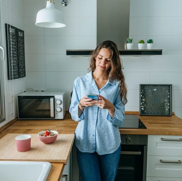 een vrouw houdt een telefoon vast en kijkt ernaar terwijl ze tegen de keuken aan staat en naast haar staat een kopje en een kommetje met eten