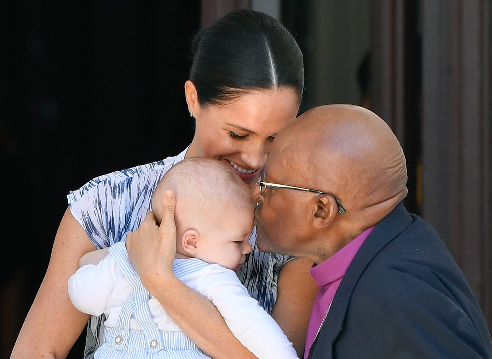 Archbishop Desmond Tutu plants a kiss on baby Archie's forehead, while Meghan Markle holds Archie with a smile.