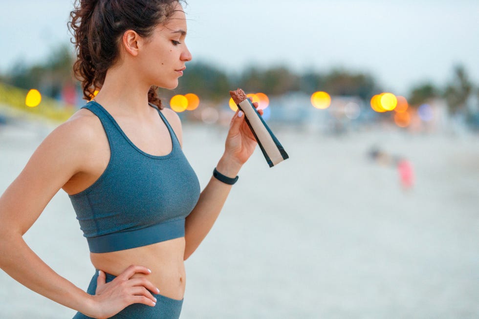 young female jogger in sports bra standing on a break and eating protein bar