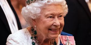 queen elizabeth ii smiles and looks right of the camera, she wears a white beaded gown and a blue sash with two pendants as well as a diamond and emerald crown and matching necklace