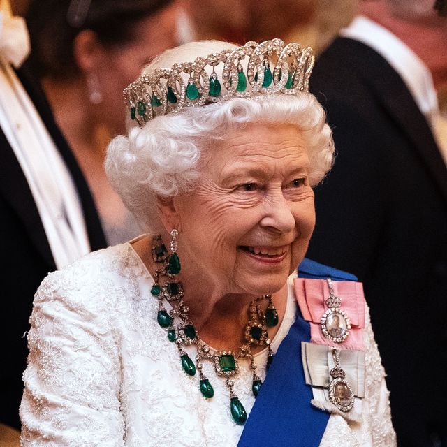 queen elizabeth ii smiles and looks right of the camera, she wears a white beaded gown and a blue sash with two pendants as well as a diamond and emerald crown and matching necklace