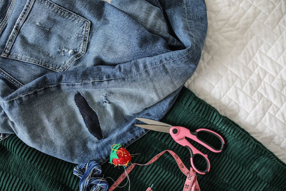 mending a pair of blue jeans with multicoloured thread, by hand scissors, measuring tape and pin cushion all lie nearby