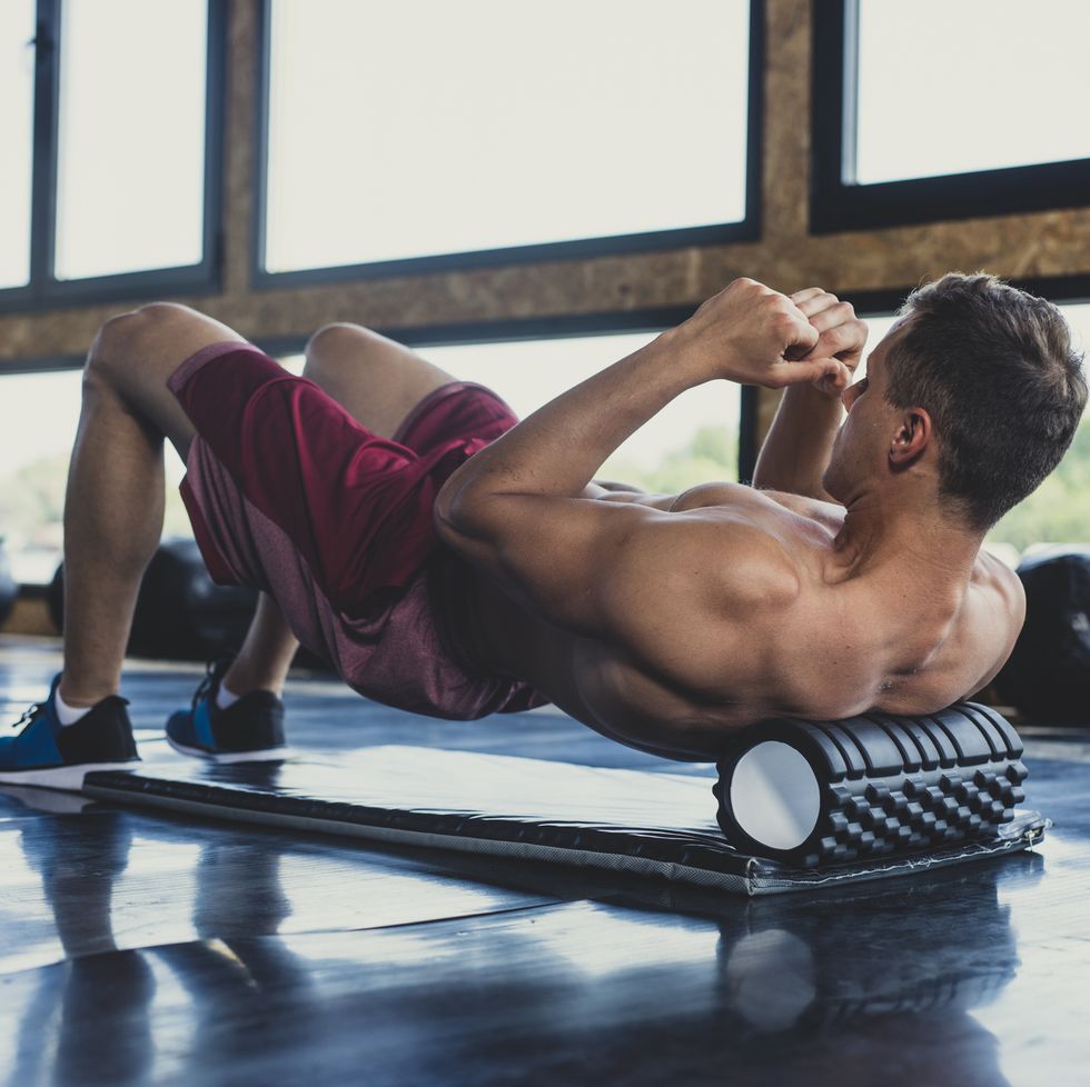 handsome young muscular caucasian man massaging his upper back with a foam roller in an indoor gym
