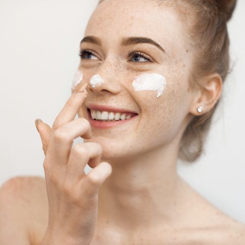 portrait of a charming young female with red hair and freckles isolated on white applying a anti age cream on her face and nose smiling