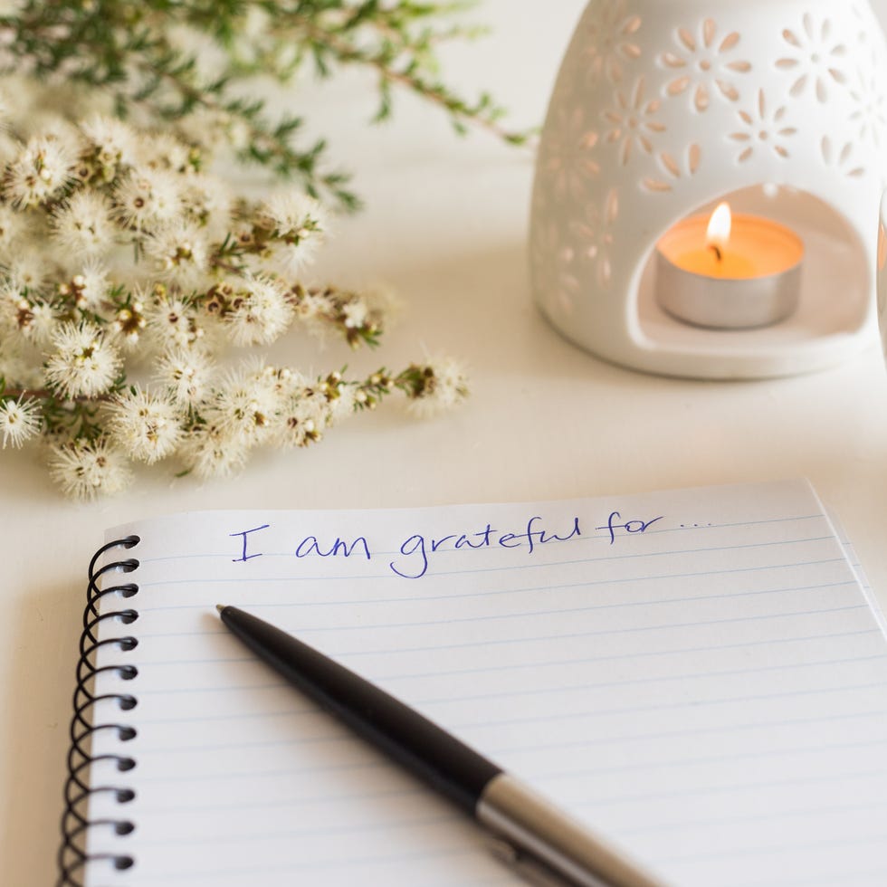 close up of handwritten text i am grateful for in foreground with notebook, pen, cup of tea, flowers and oil burner in soft focus