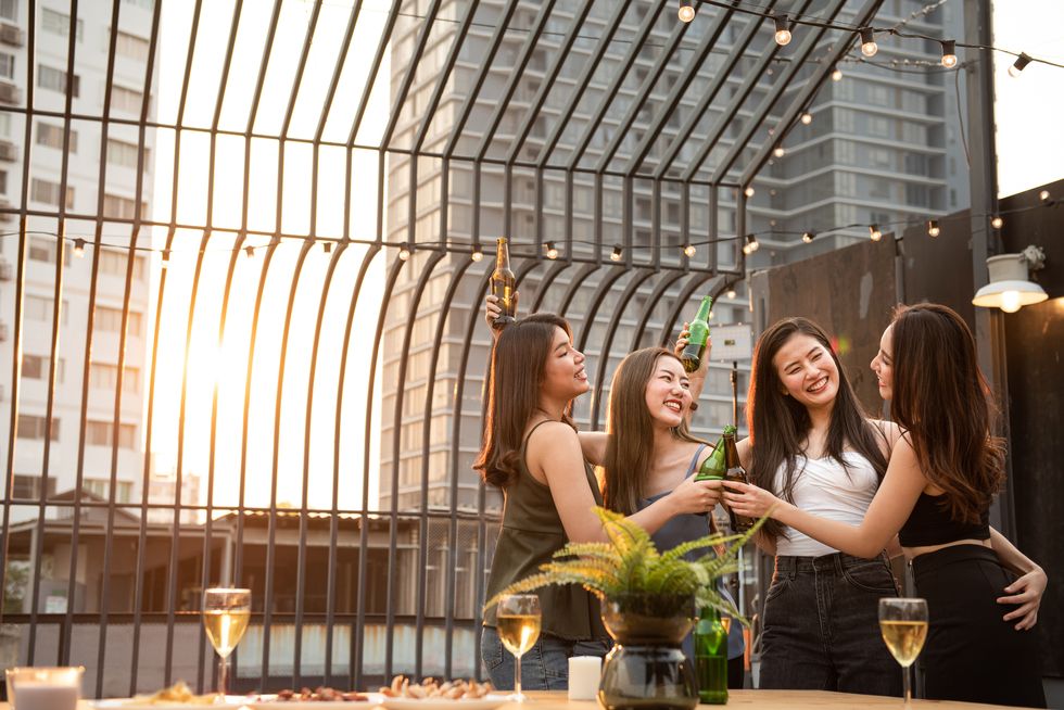 group of young beautiful happy asian women holding bottle of beer chat together with friends while celebrating dance party on outdoor rooftop nightclub with copy space for advertising