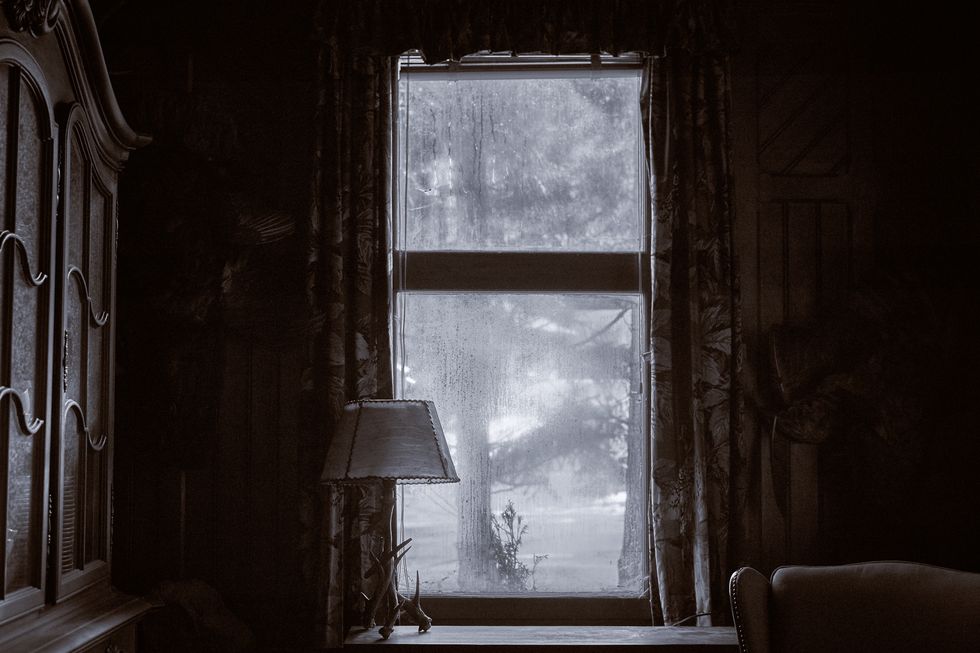 looking out of a dirty window with a curtain and a vintage lamp on the windowsill wooden living room cabinet and home interior a tree is in front of the spooky house
