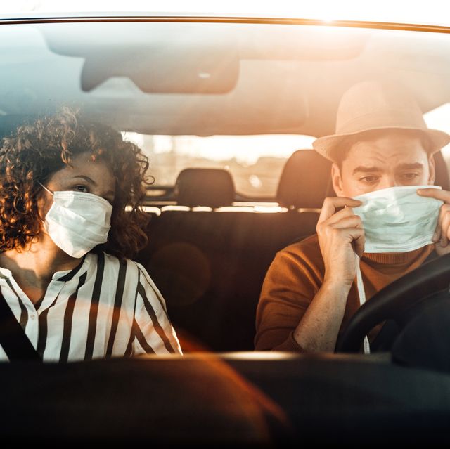 corona virus pandemic concept breathing through a medical mask because of the danger of getting the flu virus, influenza infection enjoying travel beautiful young couple sitting on the front passenger seats and smiling while handsome man driving a car