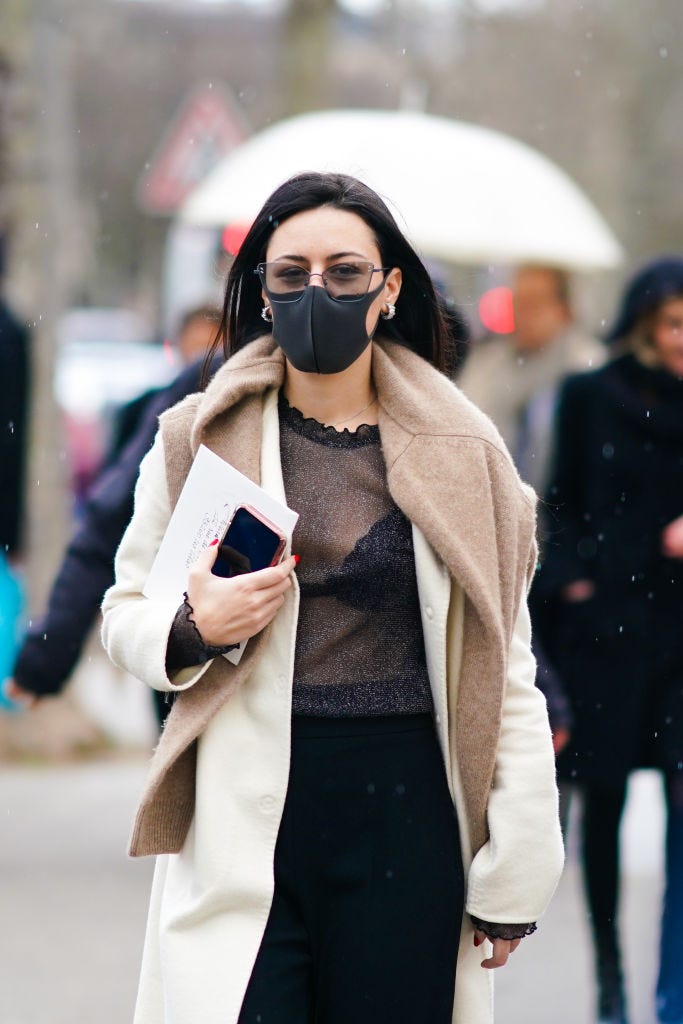 paris, france   february 29 a guest wears glasses, a brown wool pullover over the shoulders, a white long coat, a mesh black top, black bras, black pants, earrings, a protective face mask in a context of coronavirus covid 19 breakout, during paris fashion week   womenswear fallwinter 20202021, on february 29, 2020 in paris, france photo by edward berthelotgetty images