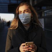 young woman standing on train station wearing protective mask, using phone