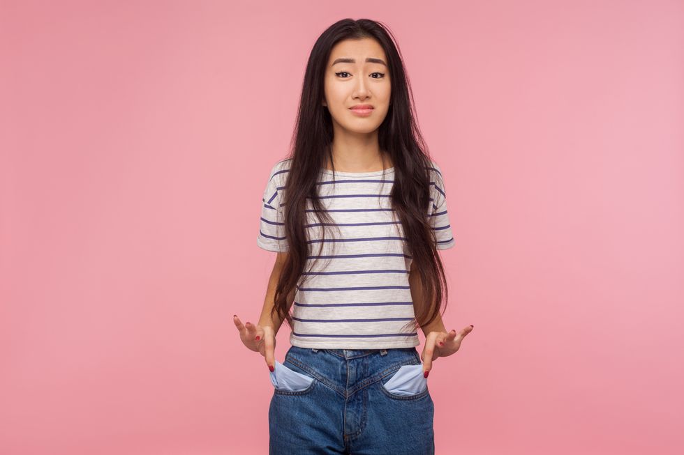 unemployment and bankruptcy upset poor girl with brunette hair in striped t shirt turning out empty pockets, worried about debts, no cash for living indoor studio shot isolated on pink background
