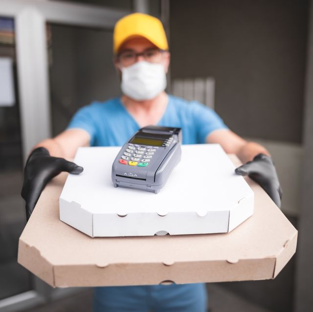 deliveryman with protective medical mask holding pizza box and pos wireless terminal for card paying   days of viruses and pandemic, food delivery to your home and safety hygiene measures