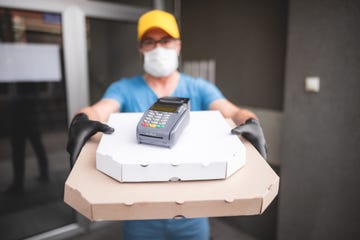 deliveryman with protective medical mask holding pizza box and pos wireless terminal for card paying   days of viruses and pandemic, food delivery to your home and safety hygiene measures