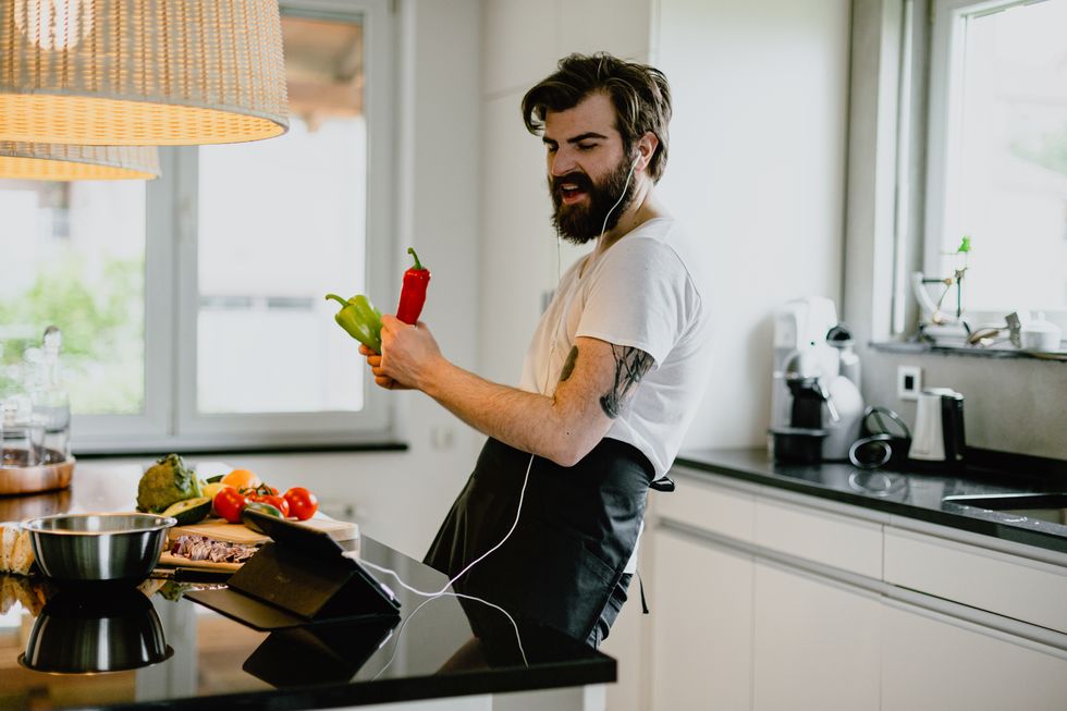 handsome bearded young man preparing healthy meal with vegetables, using headphones listening to music and singing in the kitchen at home