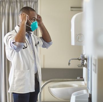 side view of teenage african male medical student wearing short sleeved lab coat and looking in mirror as he puts on n95 face mask
