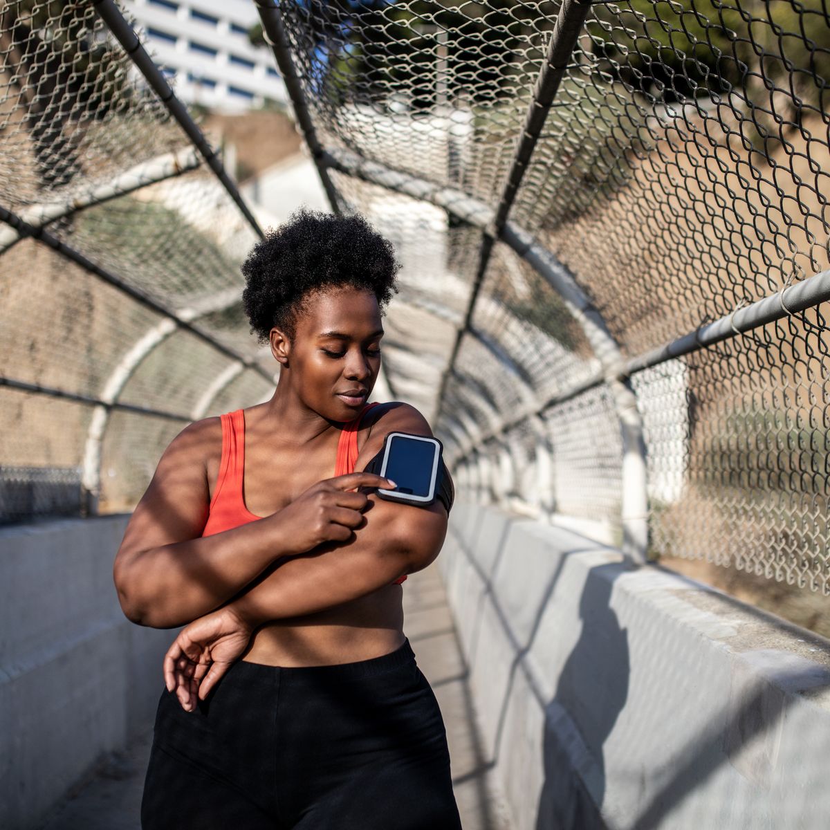 healthy woman touching phone screen on armband before exercising outdoors african american woman in sports clothing using phone while exercising outdoors