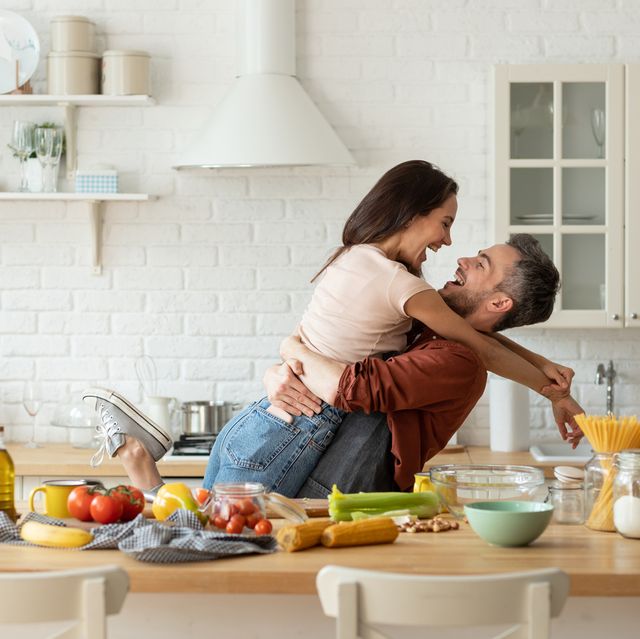 happy husband and wife having fun during cooking at home kitchen excited man hugging and carrying loving woman, cozy bright loft interior with table full of fresh healthy groceries food laughter fun
