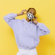 a young girl stands in front of a yellow background and wears a protective mask during the coronavirus she wears lavender hoodie and white jeans and her elbows are wide apart