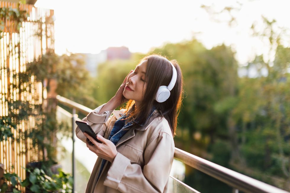 young asian woman with eyes closed enjoying music over headphones and using smartphone, relaxing on the balcony against sunlight