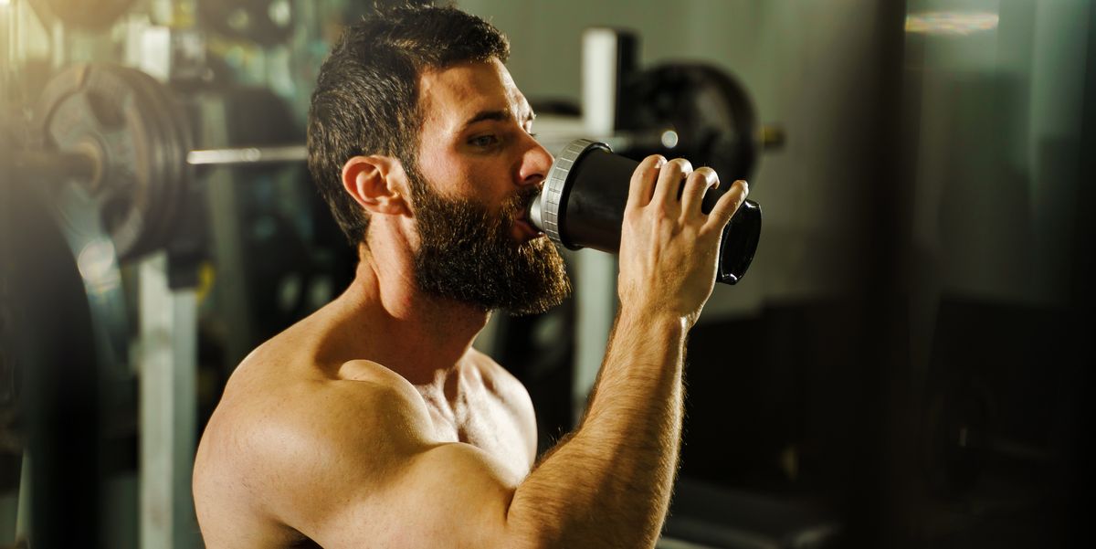 side view portrait of young muscular caucasian man bodybuilder shirtless male sitting in dark gym holding protein supplement shaker drinking supplementation in training waist up black hair and beard