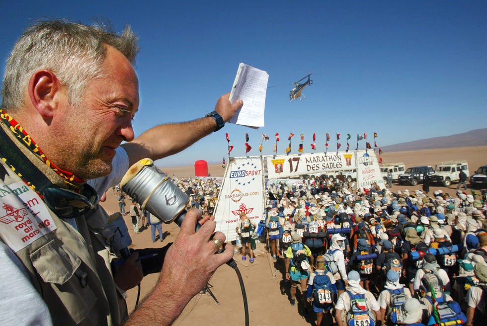 patrick bauer addressing marathon des sables competitors at the start line through a microphone and holding up his arm as a helicopter circles over ahead in the very blue sky.