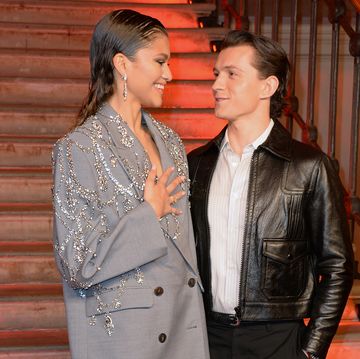 zendaya tom holland pictured smiling at one another zendaya is wearing a grey suit with silver embellishments and tom is wearing a leather jacket over a white shirt