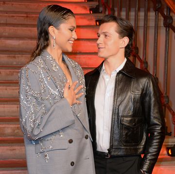 zendaya tom holland pictured smiling at one another zendaya is wearing a grey suit with silver embellishments and tom is wearing a leather jacket over a white shirt