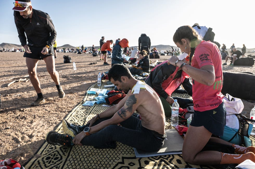 competitors rest and tend to their injuries after running part of the marathon des sables and a woman examines a man's bandaged back.