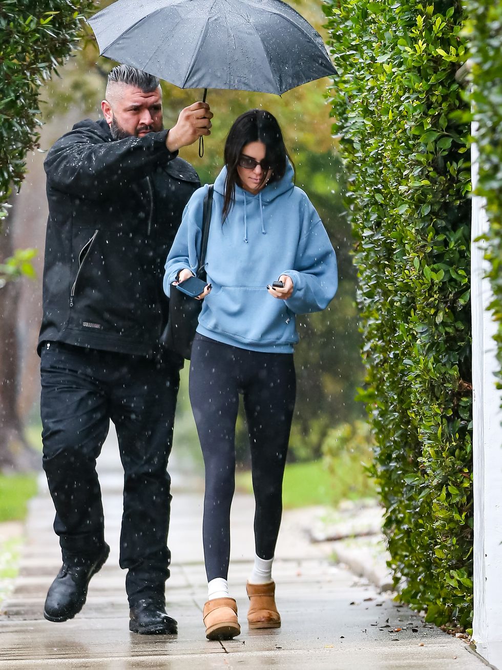 kendall jenner walks in the rain in la wearing a blue hoodie, black leggings, white socks, and ugg minis, while her bodyguard holds an umbrella over her head
