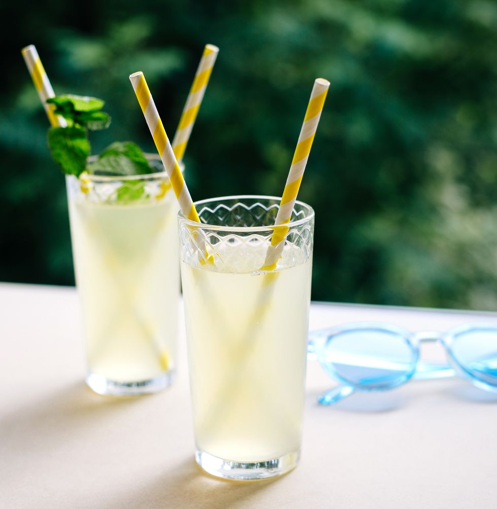 summer cold drink in glass, lemonade on table in sunny day, green trees blurred background striped yellow paper straws