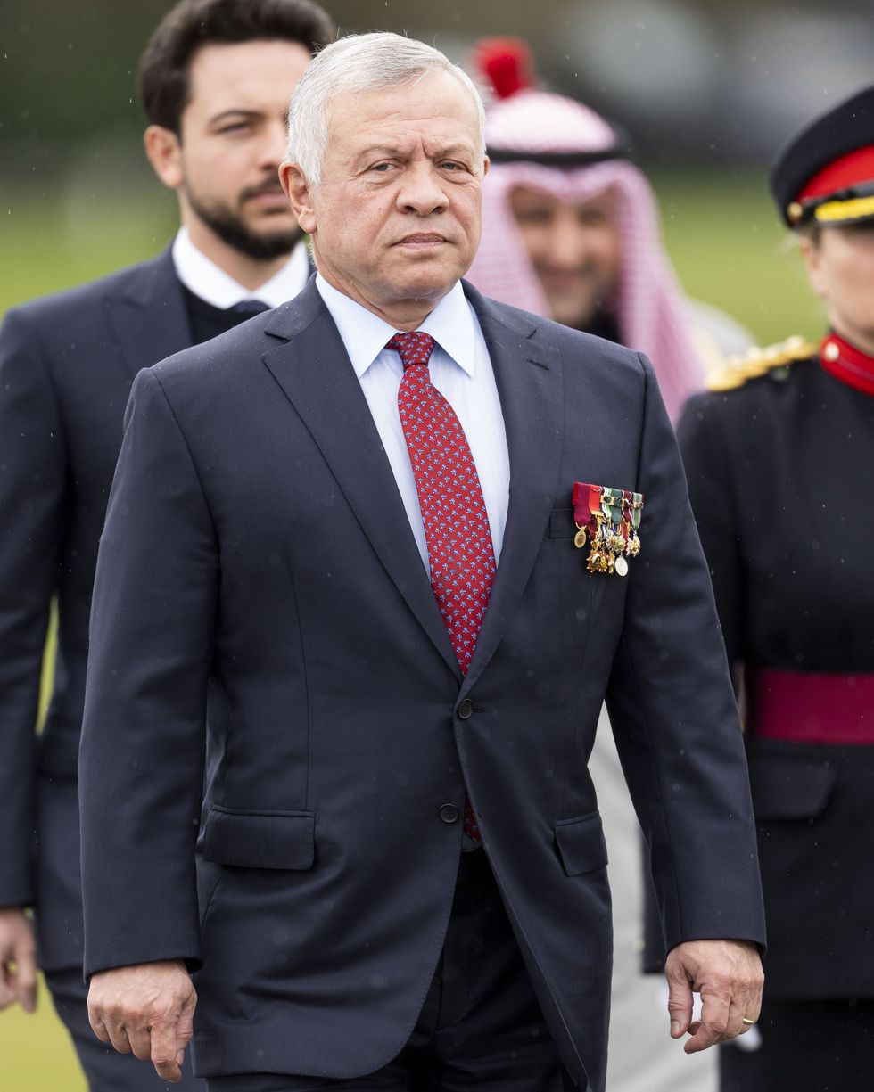 king abdullah ii of jordan stands and looks at the camera with a neutral expression, he wears a dark suit, white collared shirt and red tie with a blue pattern, his breast pocket is decorated with several medals