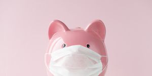 piggy bank with protective face mask on white table in front of a pink wall representing medical costs