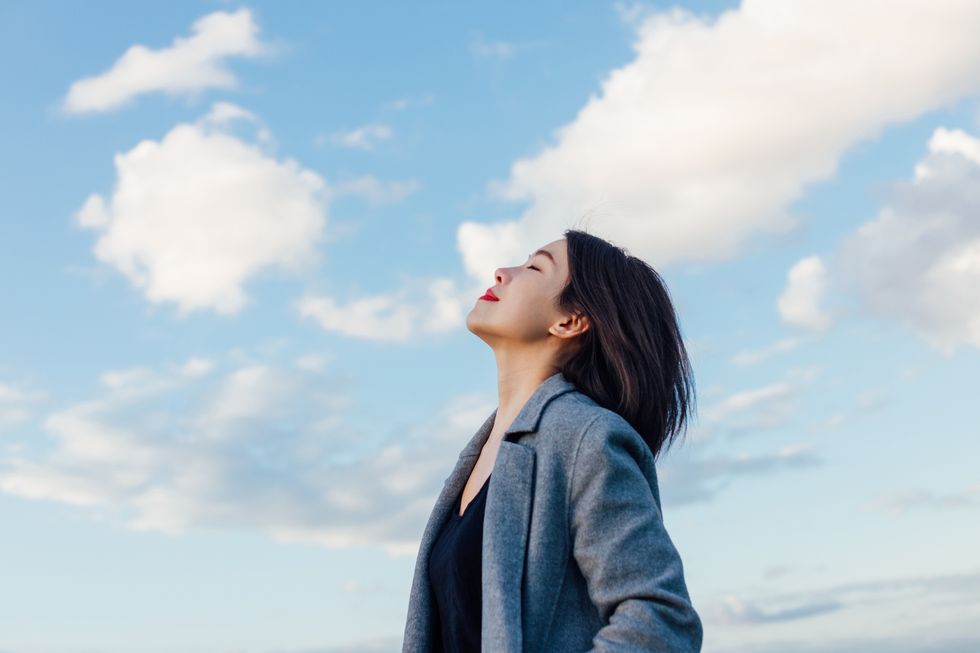 portrait of positive young asian woman with eye closed, enjoying sunlight under blue sky and clouds