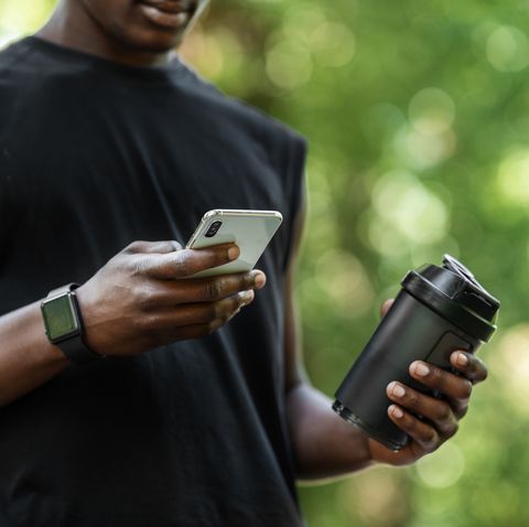 fitness mobile application cropped of african sportsman checking on sport app on mobile phone during break, holding bottle with protein drink, blurred park background, copy space