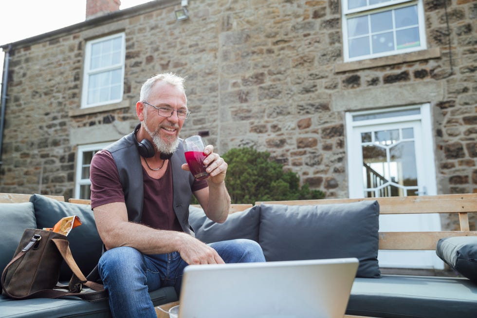 a mature caucasian man having a video call on a digital tablet while sitting outdoors on a bench with cushions he is taking a break from working from home and drinking beetroot juice out of a glass