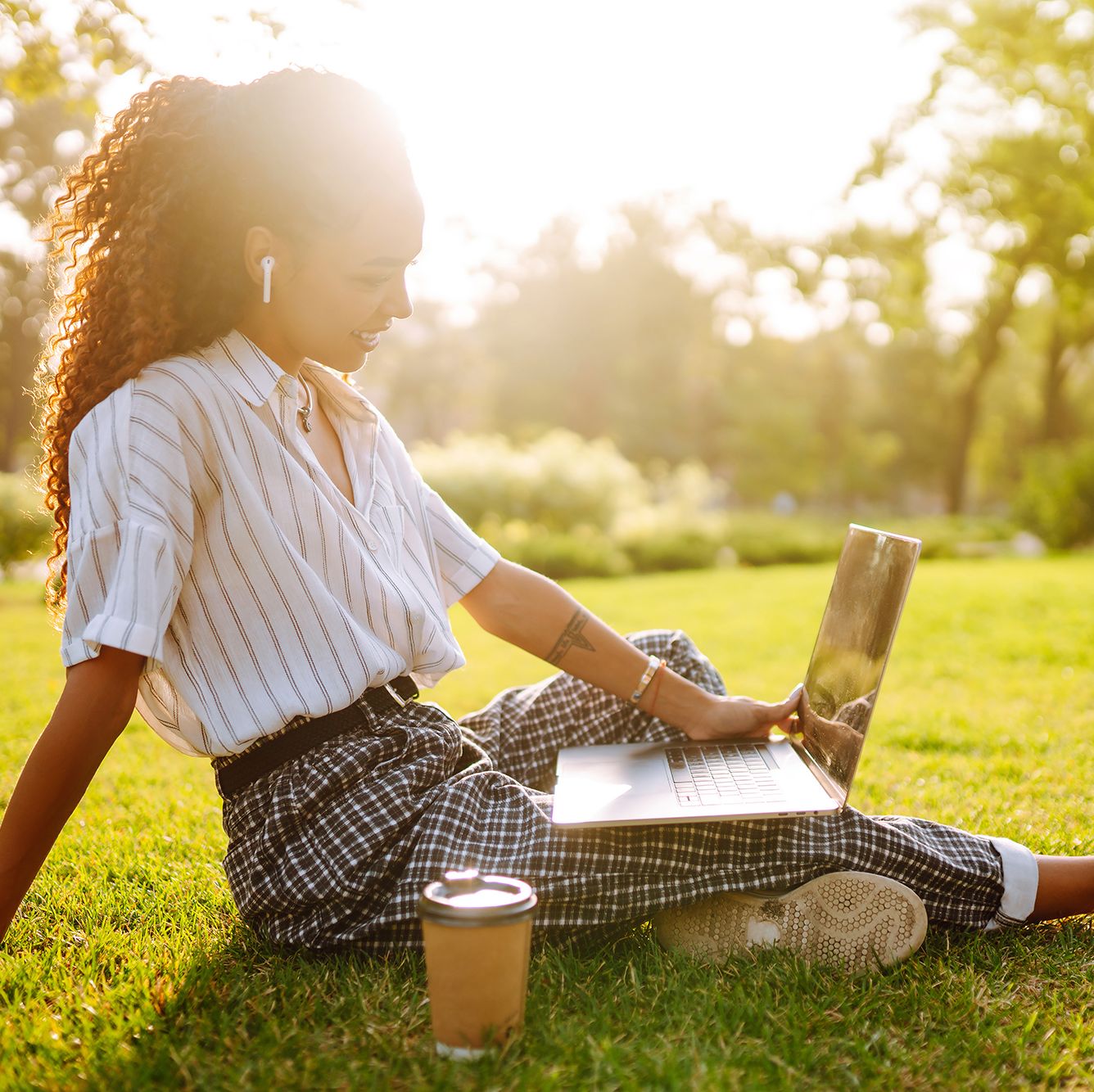 young woman sitting on green grass with laptop and coffeesmiling woman using laptop and wireless earphone for video call sunset light lifestyle concept